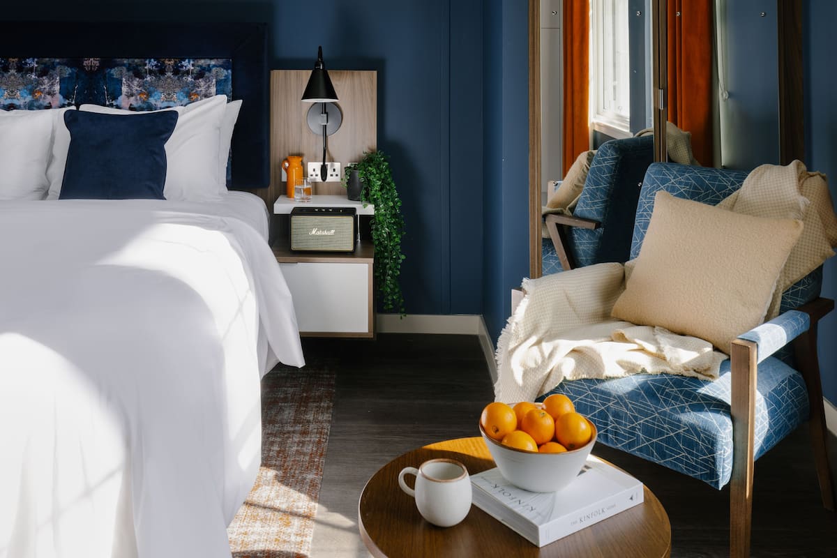 A modern hotel room with a bed, a chair, a bedside table with a lamp, a bowl of oranges, and a book.