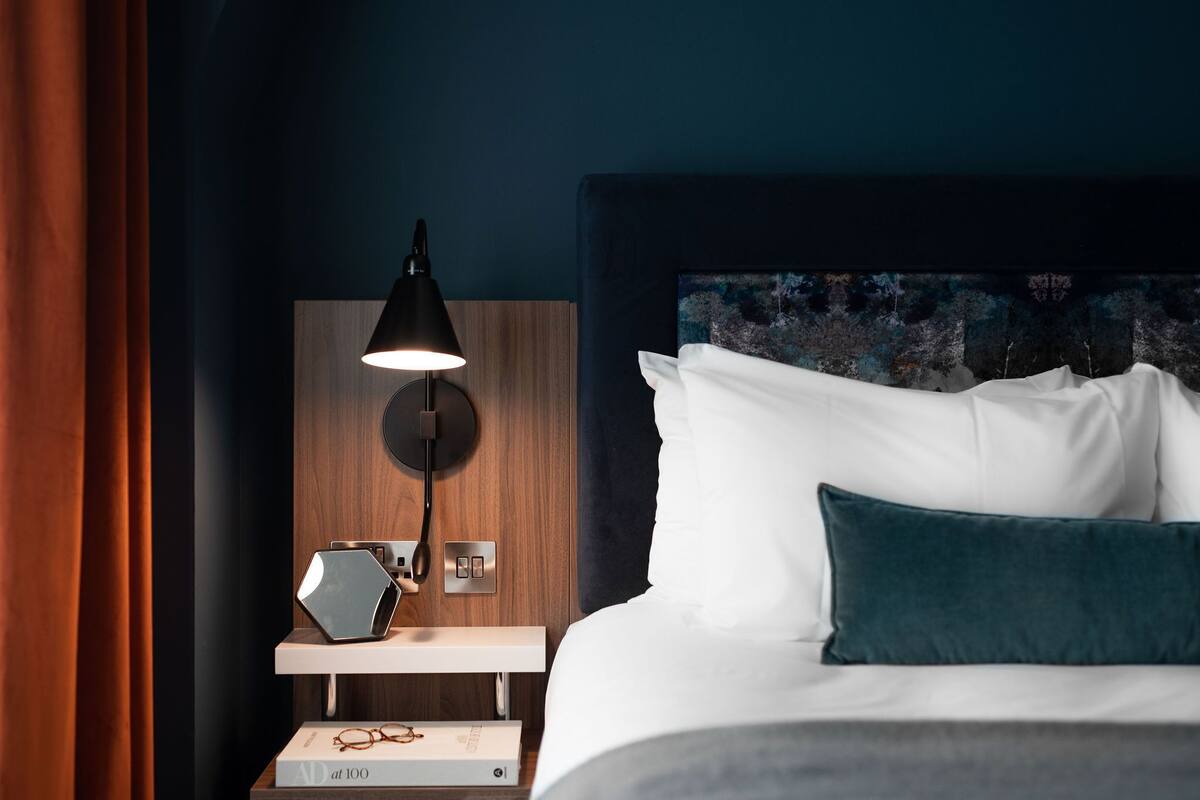 A neatly made bed with white and teal pillows, a dark headboard, and a bedside table with a lamp and a book.