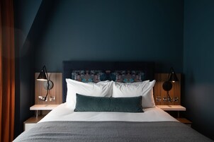 A hotel room with a large bed, two bedside tables, a headboard with a colorful artwork, and two wall-mounted lamps.