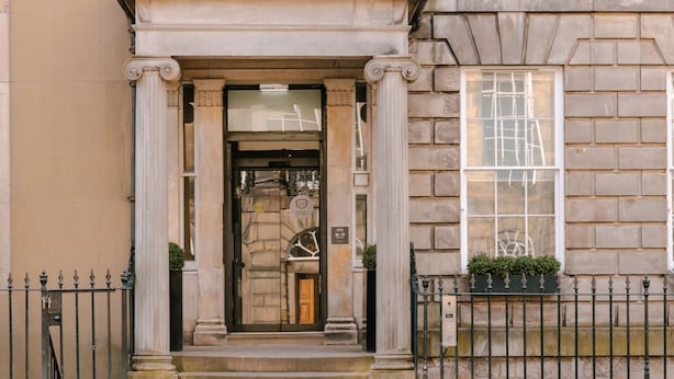 A building entrance with a classical design, featuring a central door with a glass panel, flanked by columns, and a small window with plants.