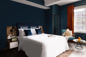 Une chambre moderne avec un grand lit, une table de chevet avec une radio, une chaise, une petite table avec un livre et une fenêtre avec des rideaux.