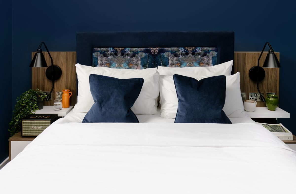 A neatly made bed with white and navy blue bedding, a wooden headboard with a colorful abstract painting, and two bedside tables with lamps and a radio.