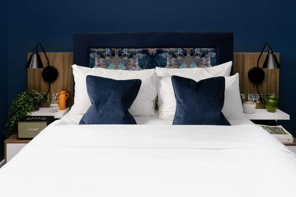 A neatly made bed with white and navy blue bedding, a wooden headboard with a colorful abstract painting, and two bedside tables with lamps and a radio.