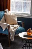 A room with a blue patterned chair, a white throw blanket, a small round table with a book and a vase, and a window with curtains.