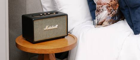 A bed with white bedding, a patterned pillow, and a Marshall speaker on a wooden side table.