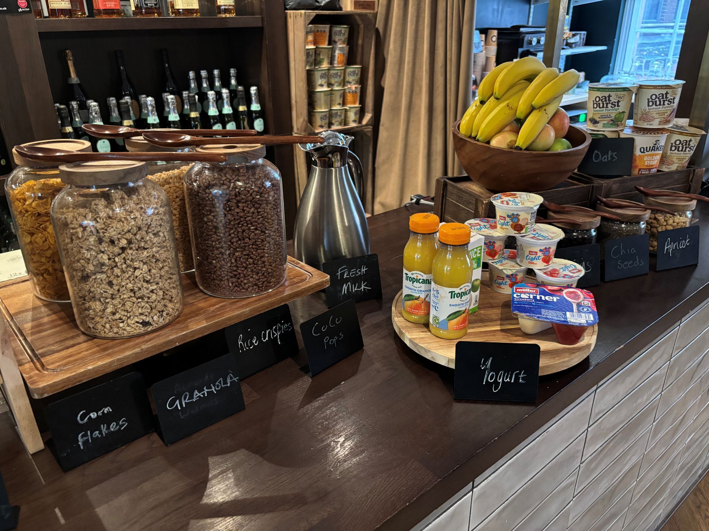 Un buffet pour le petit-déjeuner proposant divers aliments et boissons, notamment des pots de granola, un saladier de bananes, ainsi que des contenants étiquetés contenant du yaourt et du jus.