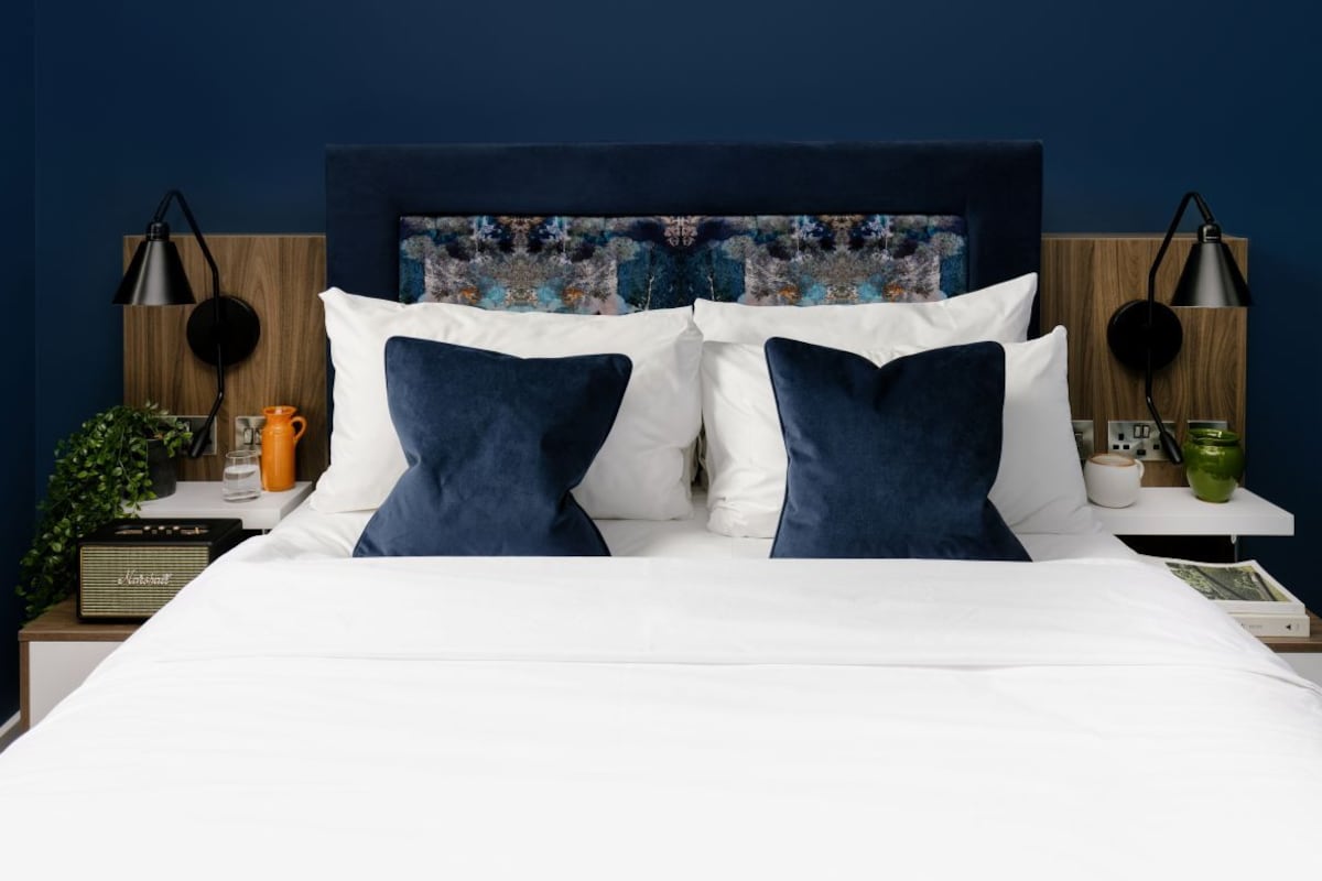 A neatly made bed with white and blue pillows, a wooden headboard with a colorful abstract painting, and two bedside tables with lamps and a radio.