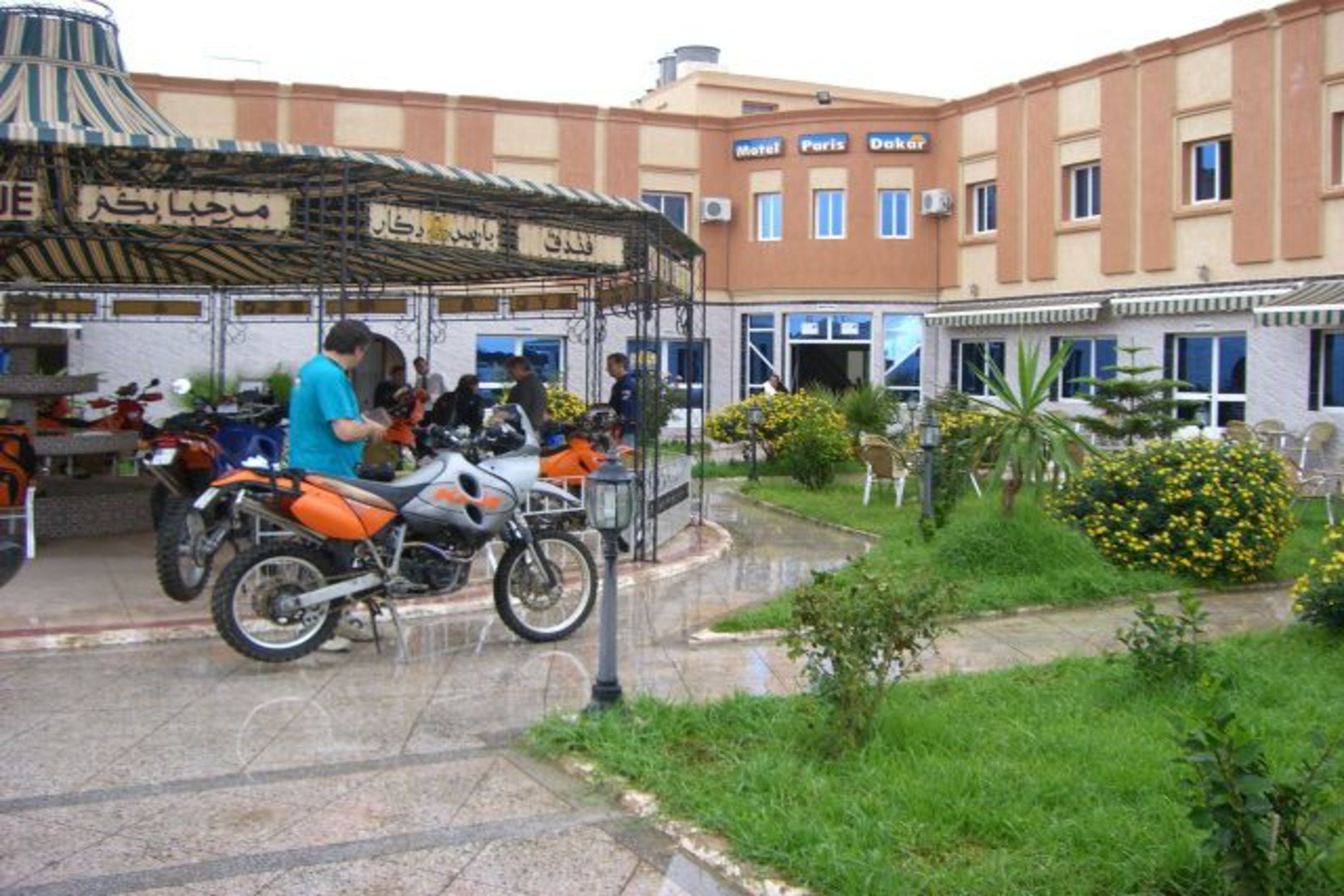 A courtyard with a covered seating area, a motorcycle, and a building with a sign that reads "Motel Paris Dakar".