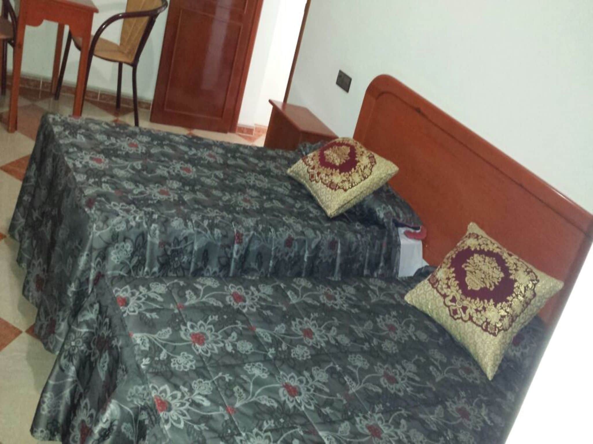 Ein Zimmer mit zwei Betten, jedes mit einem gemusterten Überwurf und dekorativen Kissen. In der Ecke steht ein Holzhocker und ein kleiner Tisch.