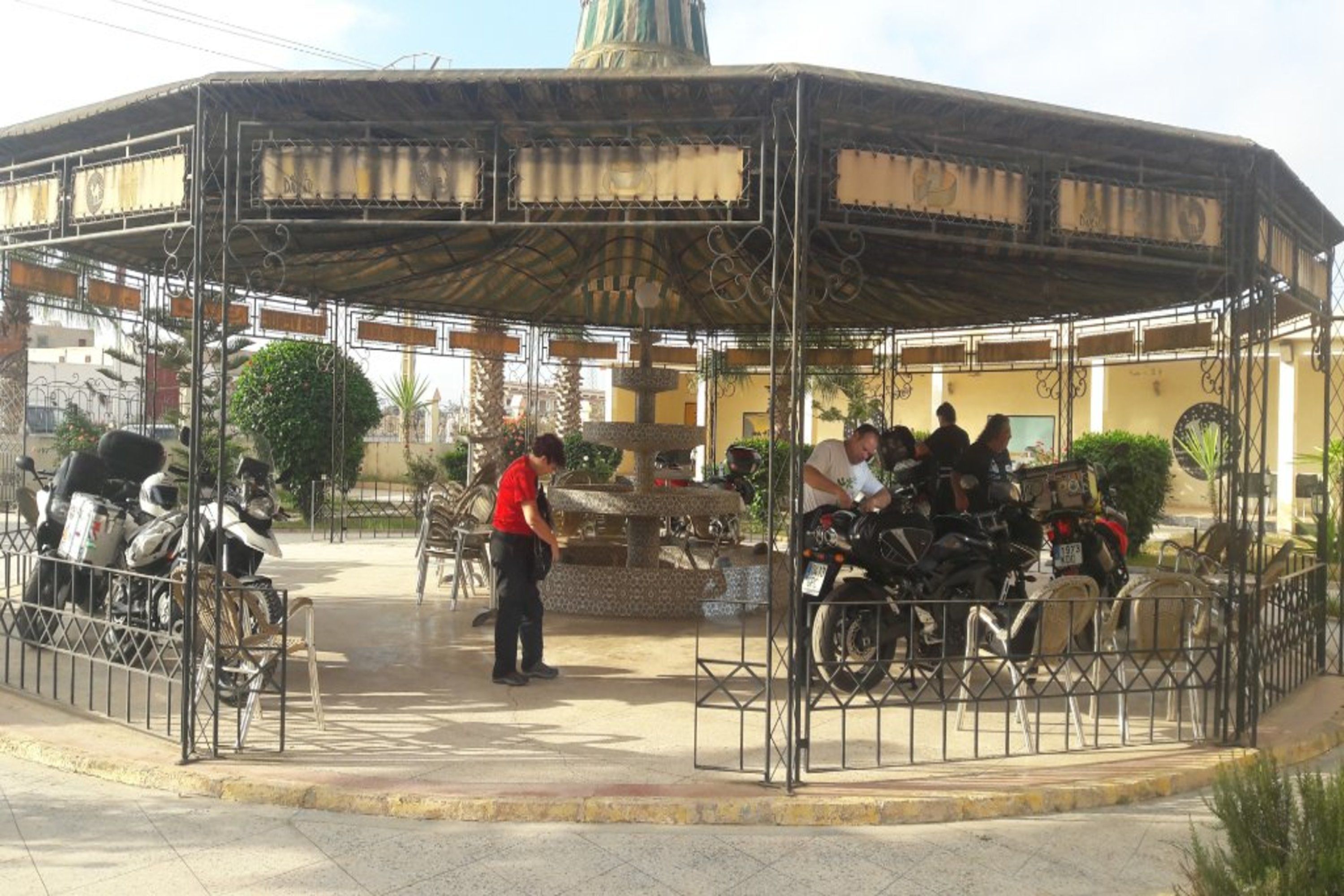 Un pabellón cubierto con una fuente central, rodeado de barandillas de metal y motocicletas estacionadas.