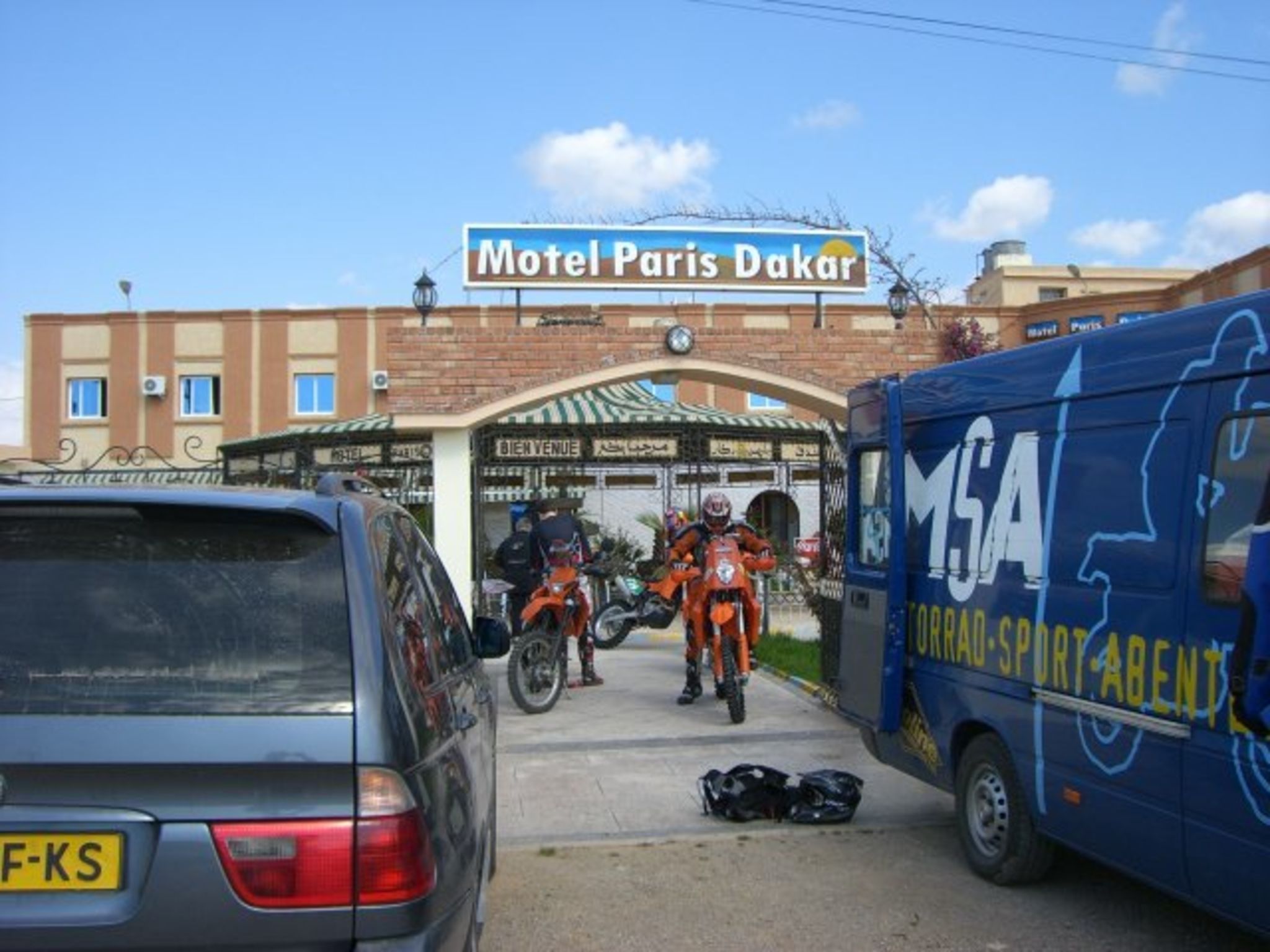 A building with a sign reading "Motel Paris Dakar" and a blue van with "MGA" and "SPORT-A-BENT" on it.