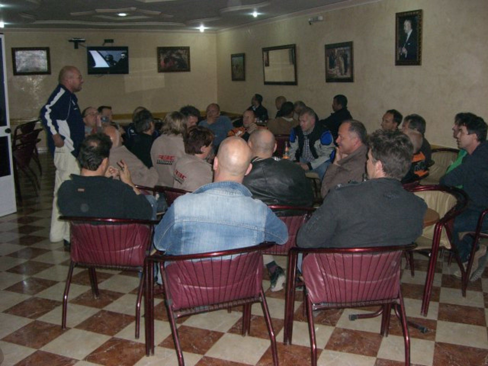 Uma sala de conferências com piso xadrez, cadeiras de metal e uma televisão na parede.