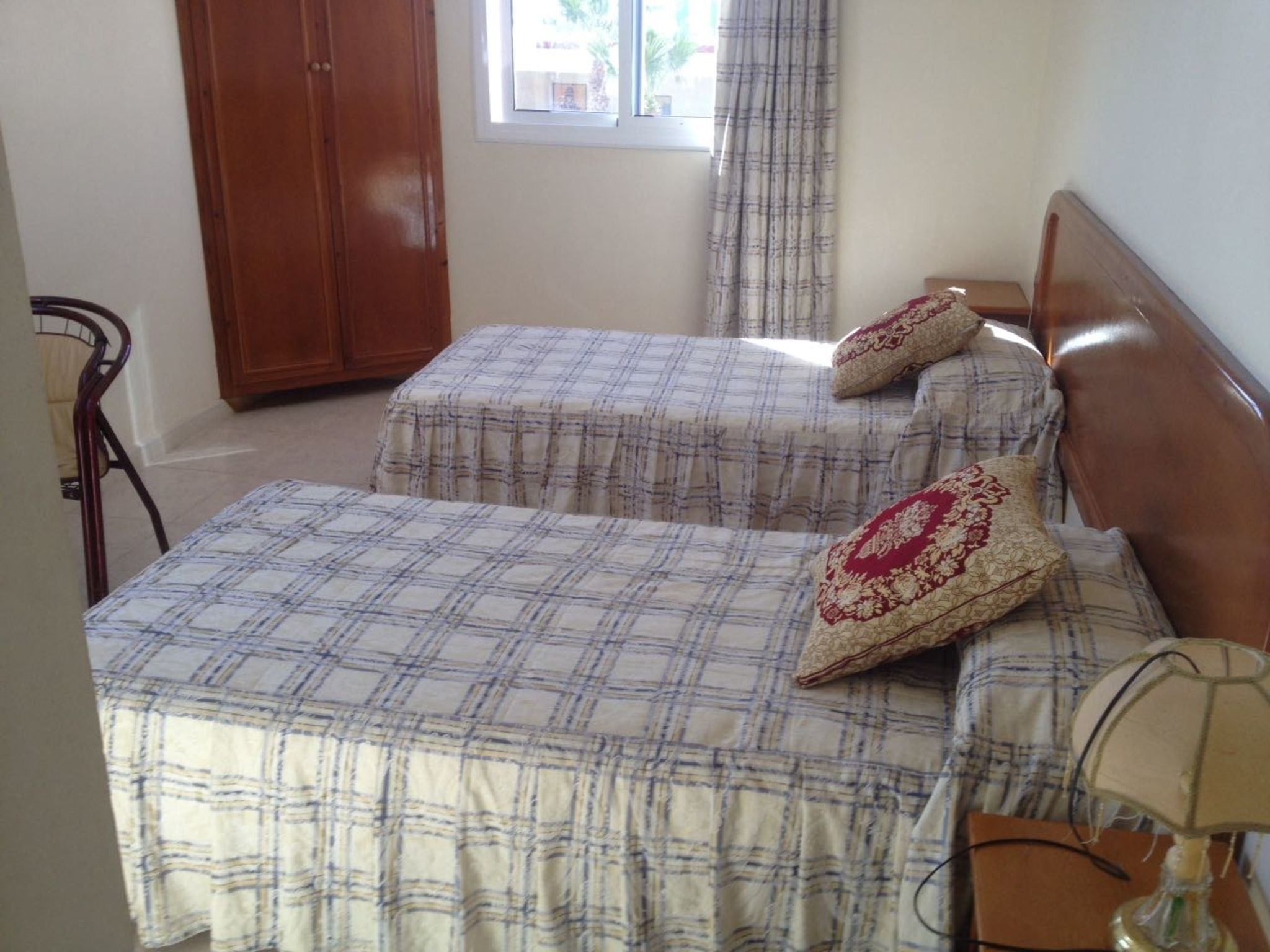 Een kamer met twee bedden, elk met een geruite plaid en decoratieve kussens. Er is een houten garderobe en een raam met gordijnen.