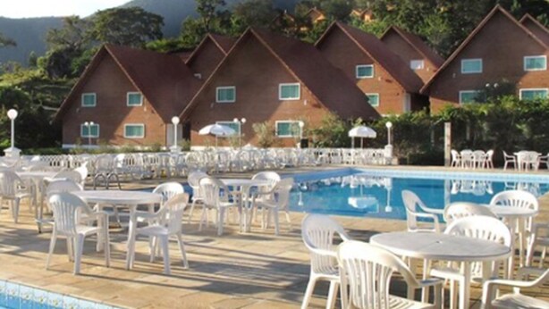 A poolside area with white plastic chairs and tables, a swimming pool, and a row of brick buildings with green windows in the background. - Resort Monte das Oliveiras (Joanópolis)