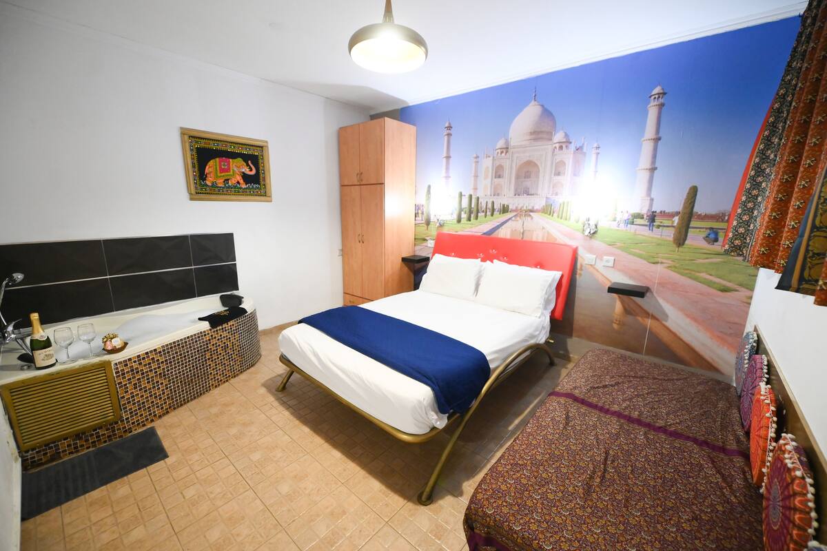 Quarto de hotel com uma cama, um guarda-roupa de madeira, uma mesinha com uma garrafa e copos, e um mural na parede com o Taj Mahal.