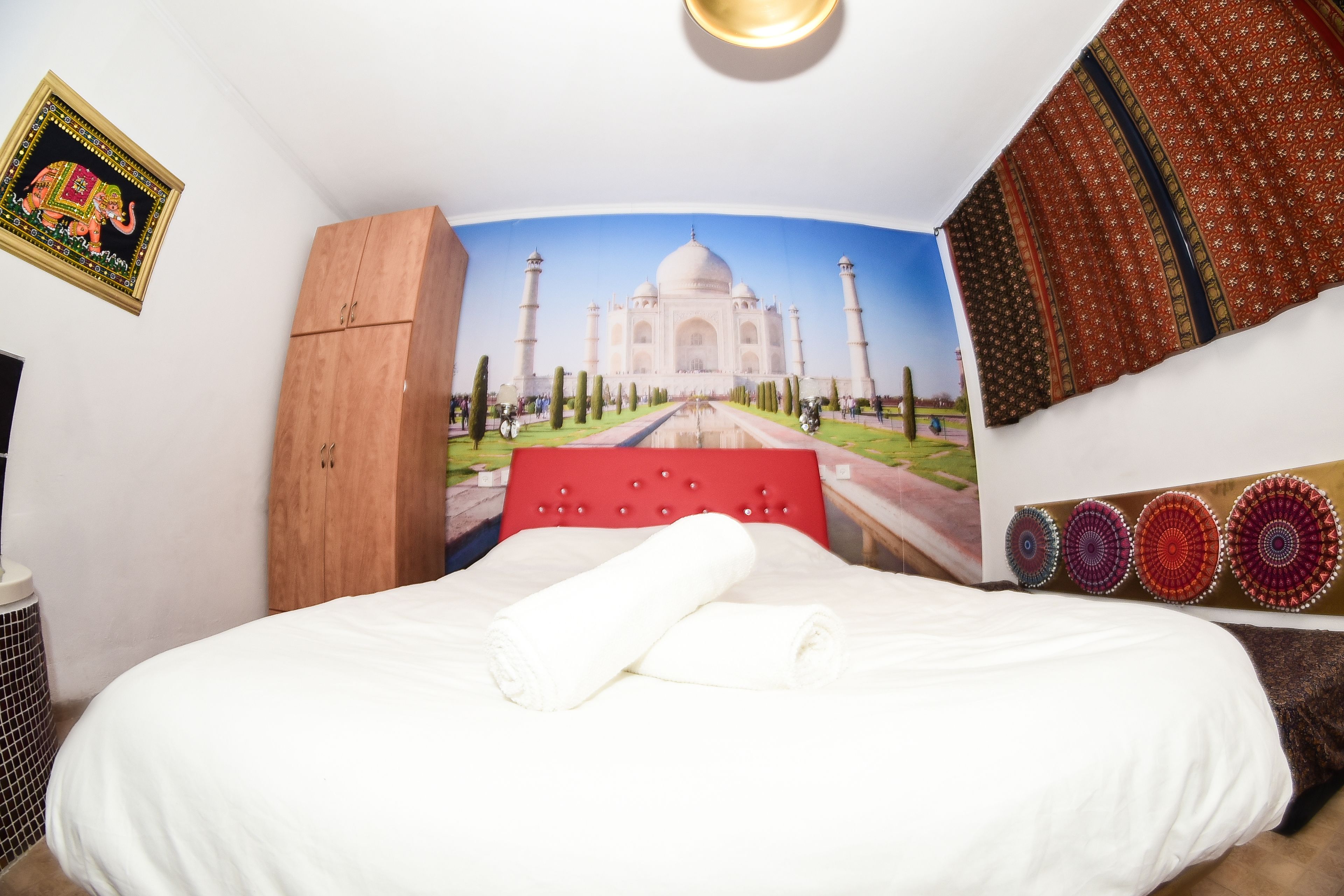 A room with a bed, a wooden wardrobe, a red headboard, and a large wall mural of the Taj Mahal.