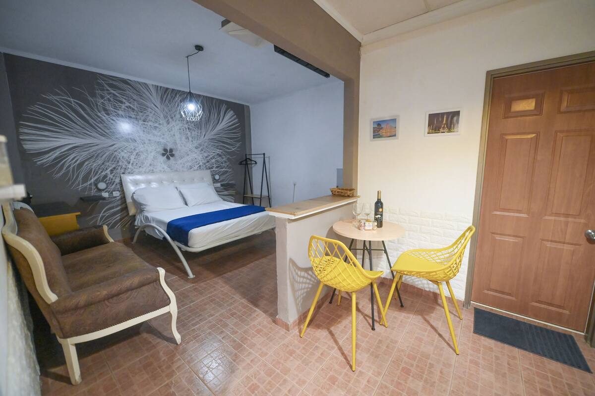 Quarto com uma cama, mesa de cabeceira com uma garrafa, duas cadeiras e uma pequena mesa.