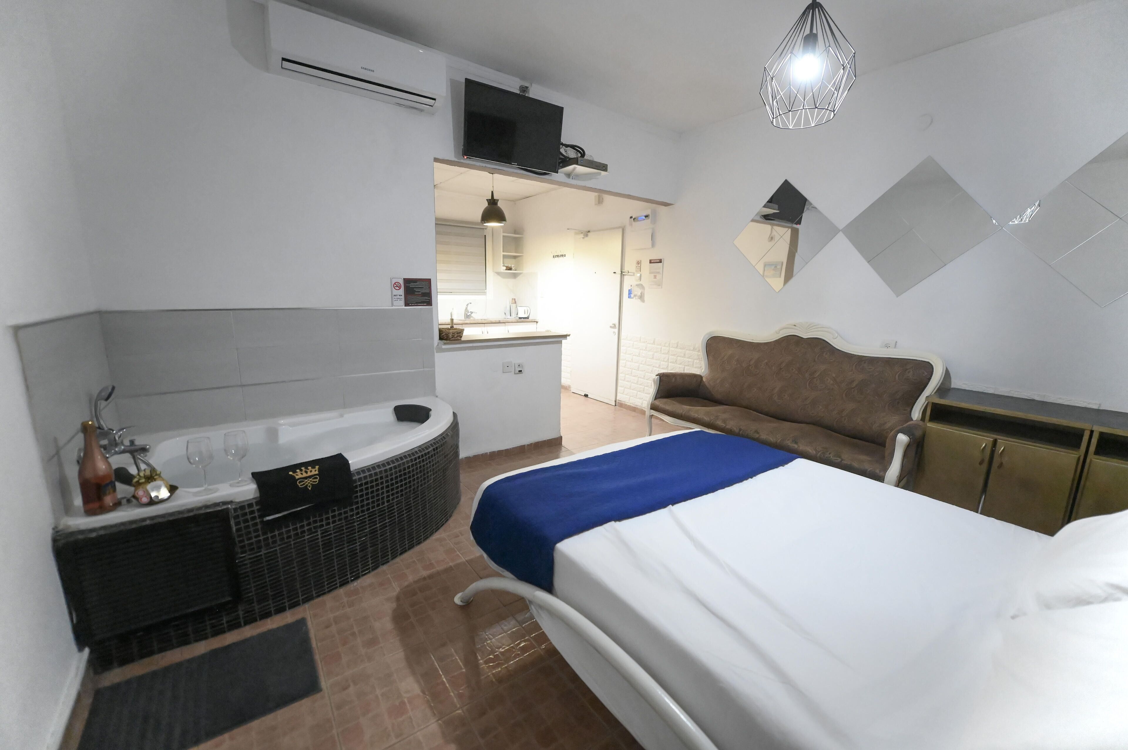 A hotel room with a bed, a sofa, a bathtub, a TV, and a kitchenette.