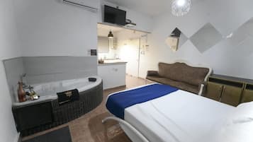 A hotel room with a bed, a sofa, a bathtub, a TV, and a kitchenette.