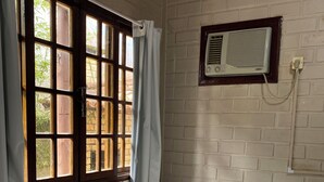 Une pièce avec un lit, une fenêtre avec des rideaux, un climatiseur fixé au mur et un mur en briques.
