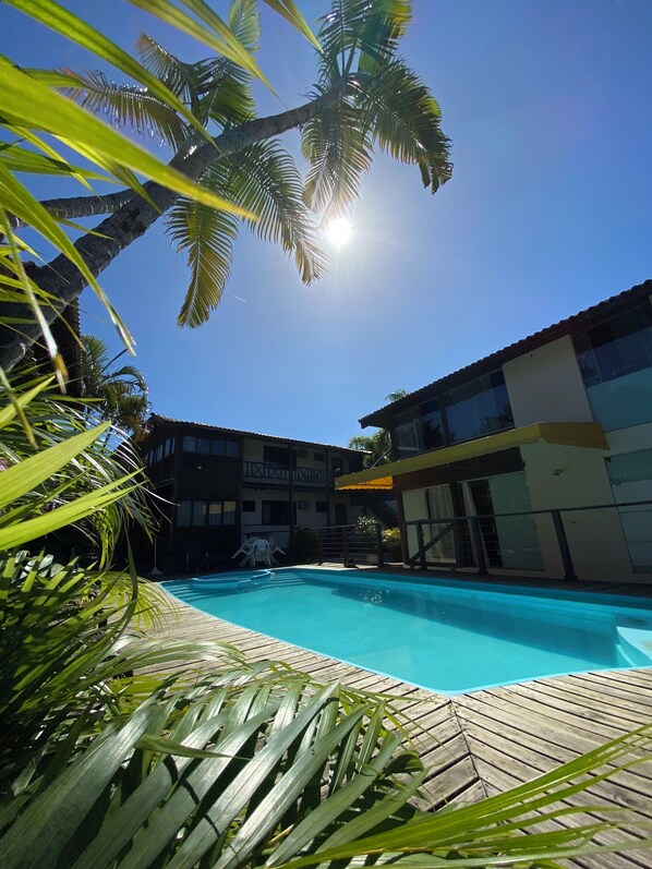 A swimming pool with a wooden deck, surrounded by palm trees and a building with balconies.