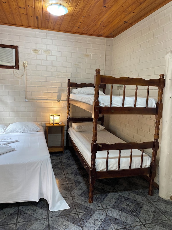 Une chambre avec un lit superposé en bois, un lit simple, une table de chevet avec une lampe et un mur en briques blanches.