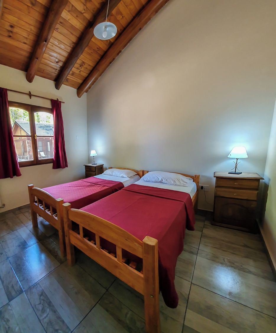 A room with two wooden beds, a window with curtains, a wooden ceiling, and a nightstand with a lamp.