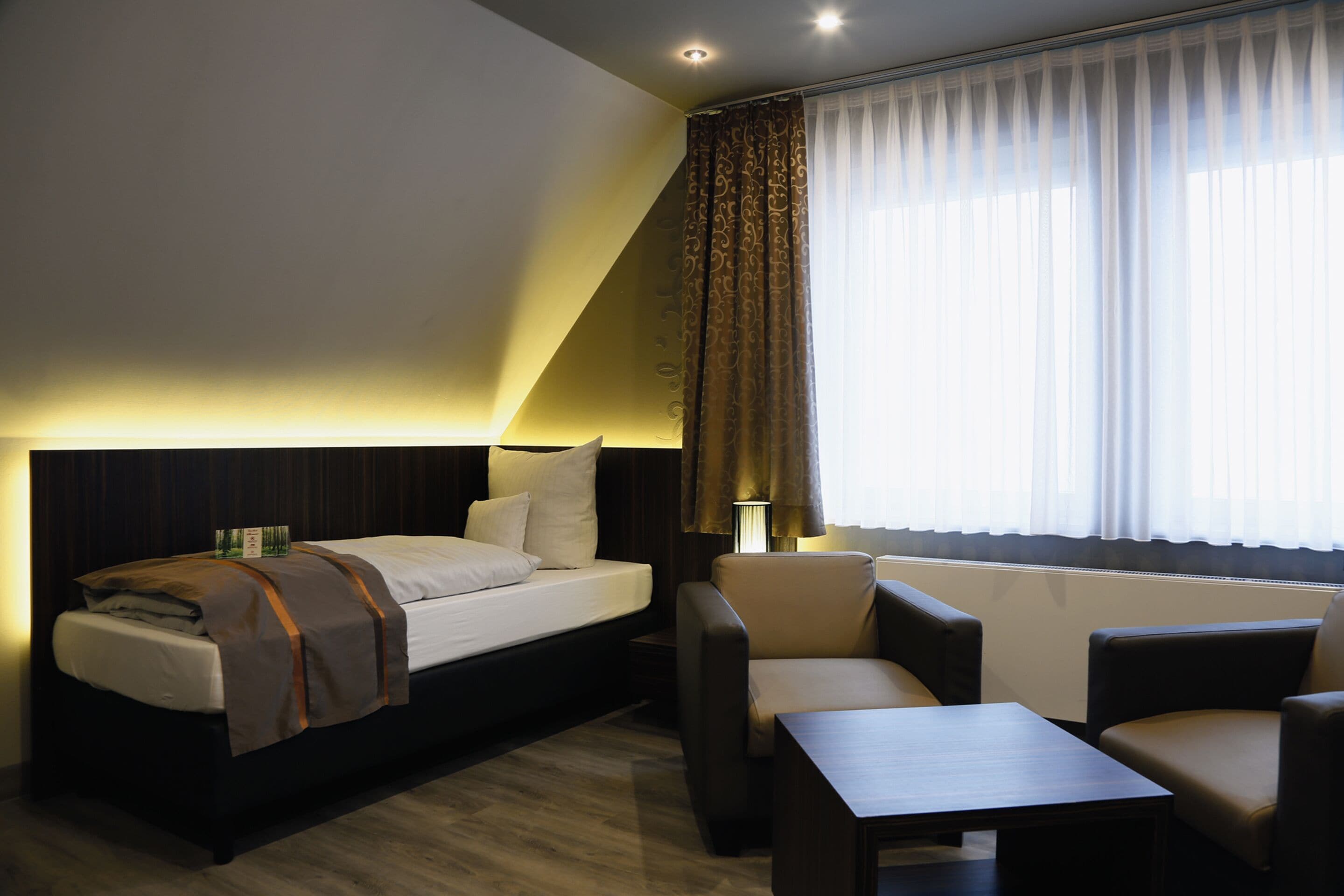 A hotel room with a bed, a desk, two chairs, a lamp, and a window with curtains.