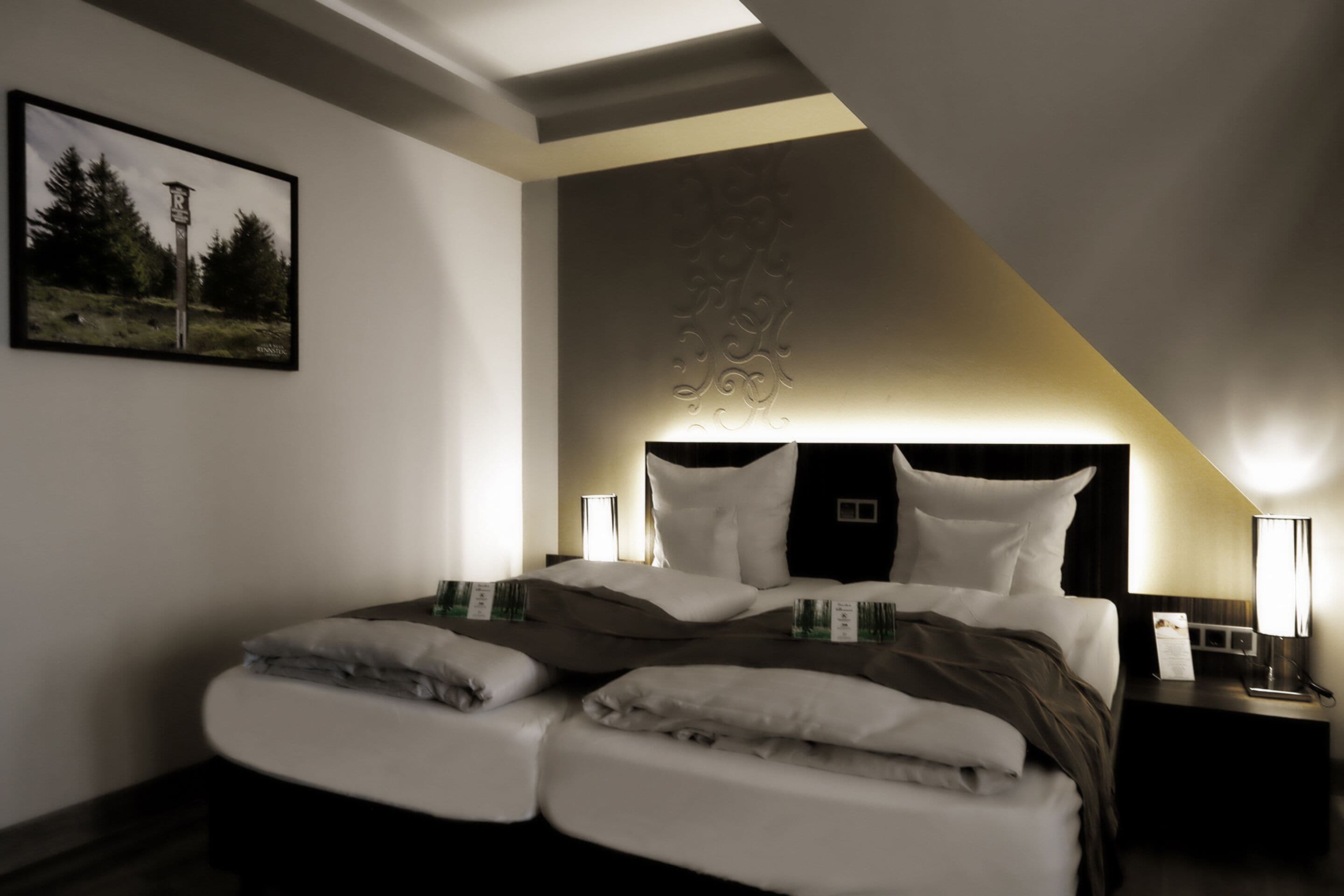 Een net opgemaakt bed met wit en bruin beddengoed, een ingelijst beeld op de muur en een nachtkastje met een lamp.