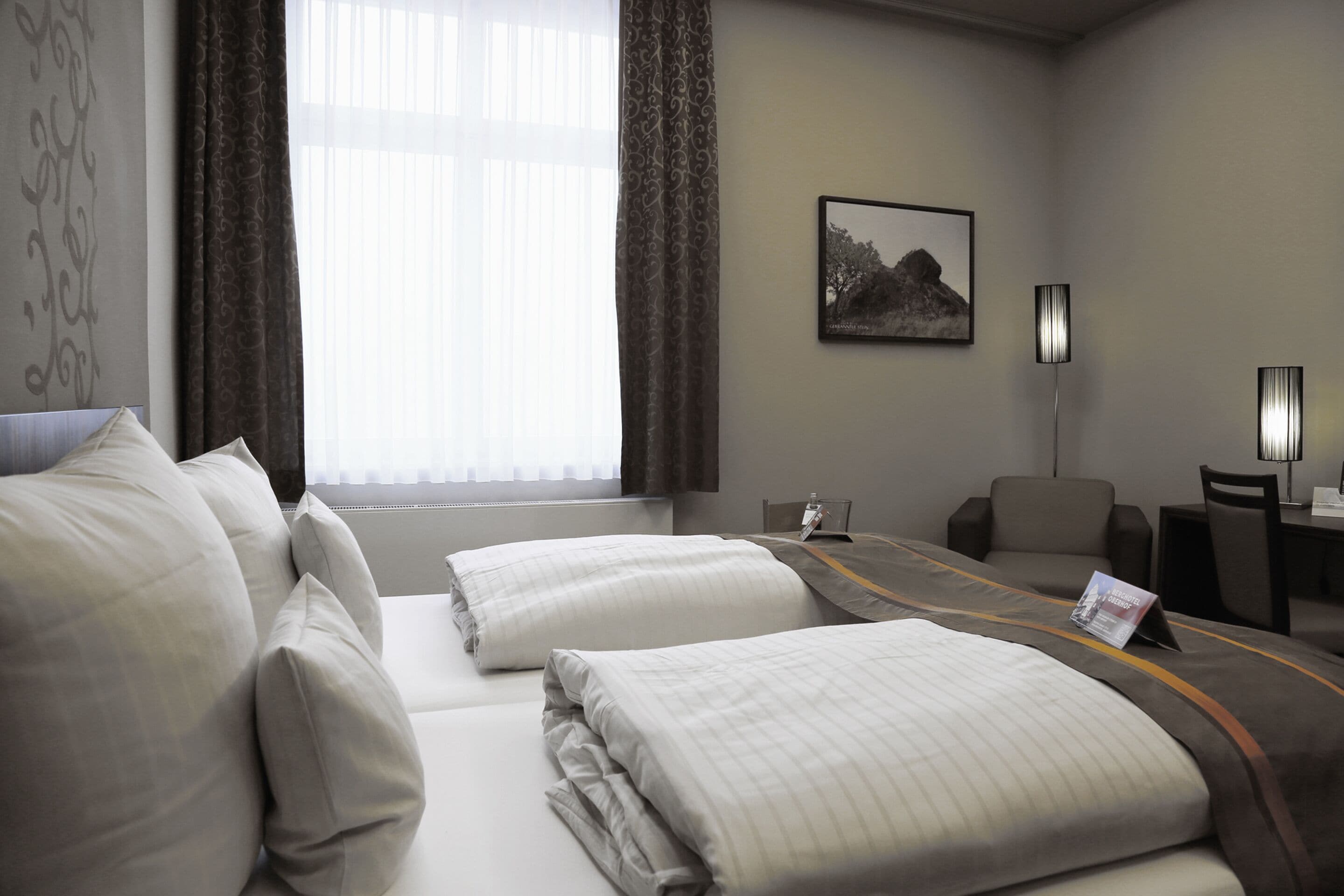 Ein Hotelzimmer mit zwei Betten, einem Schreibtisch, einem Stuhl, einem gerahmten Bild an der Wand und einem Fenster mit Vorhängen.