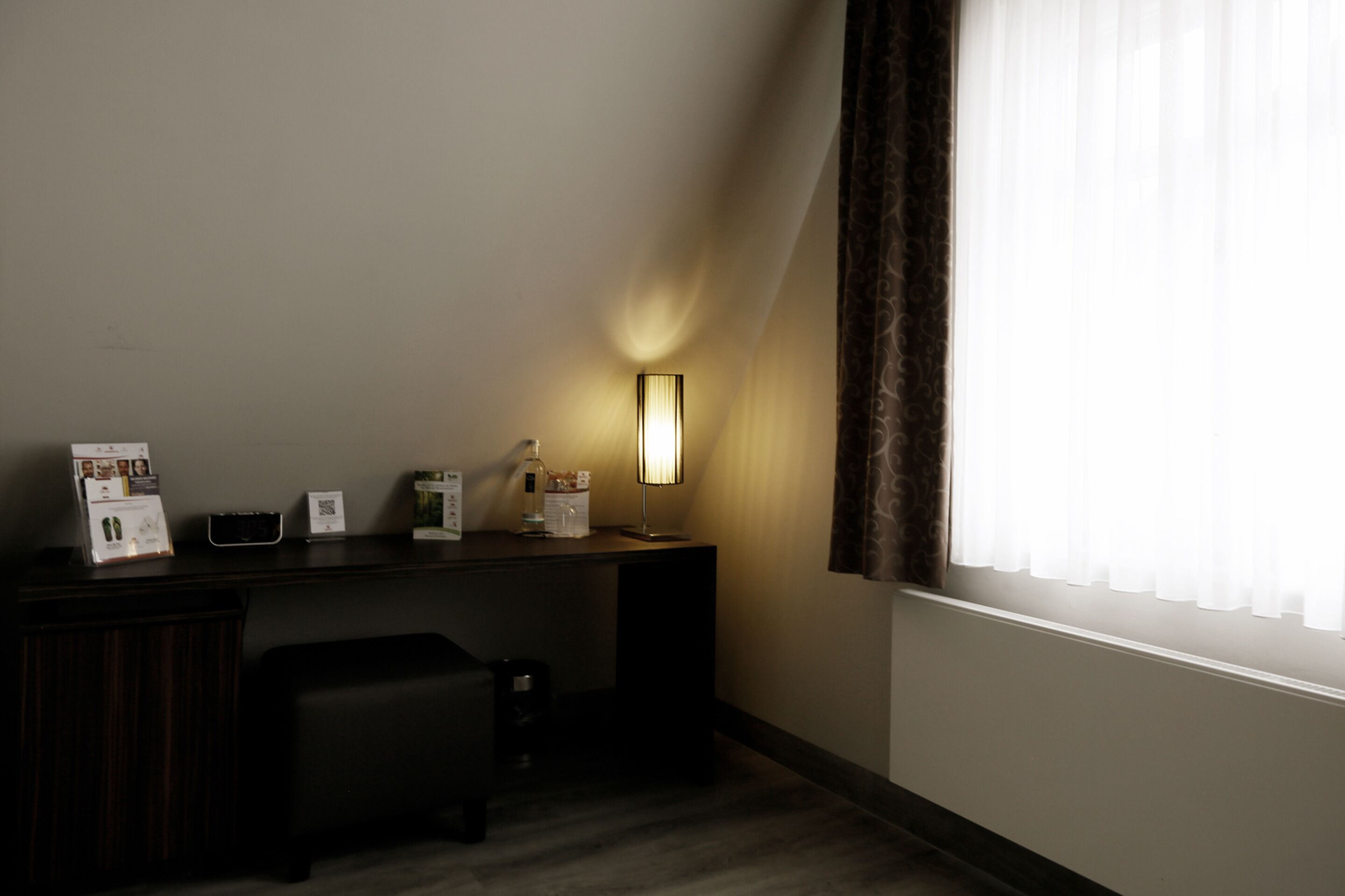 A hotel room with a desk, a lamp, a small table, and a window with curtains.