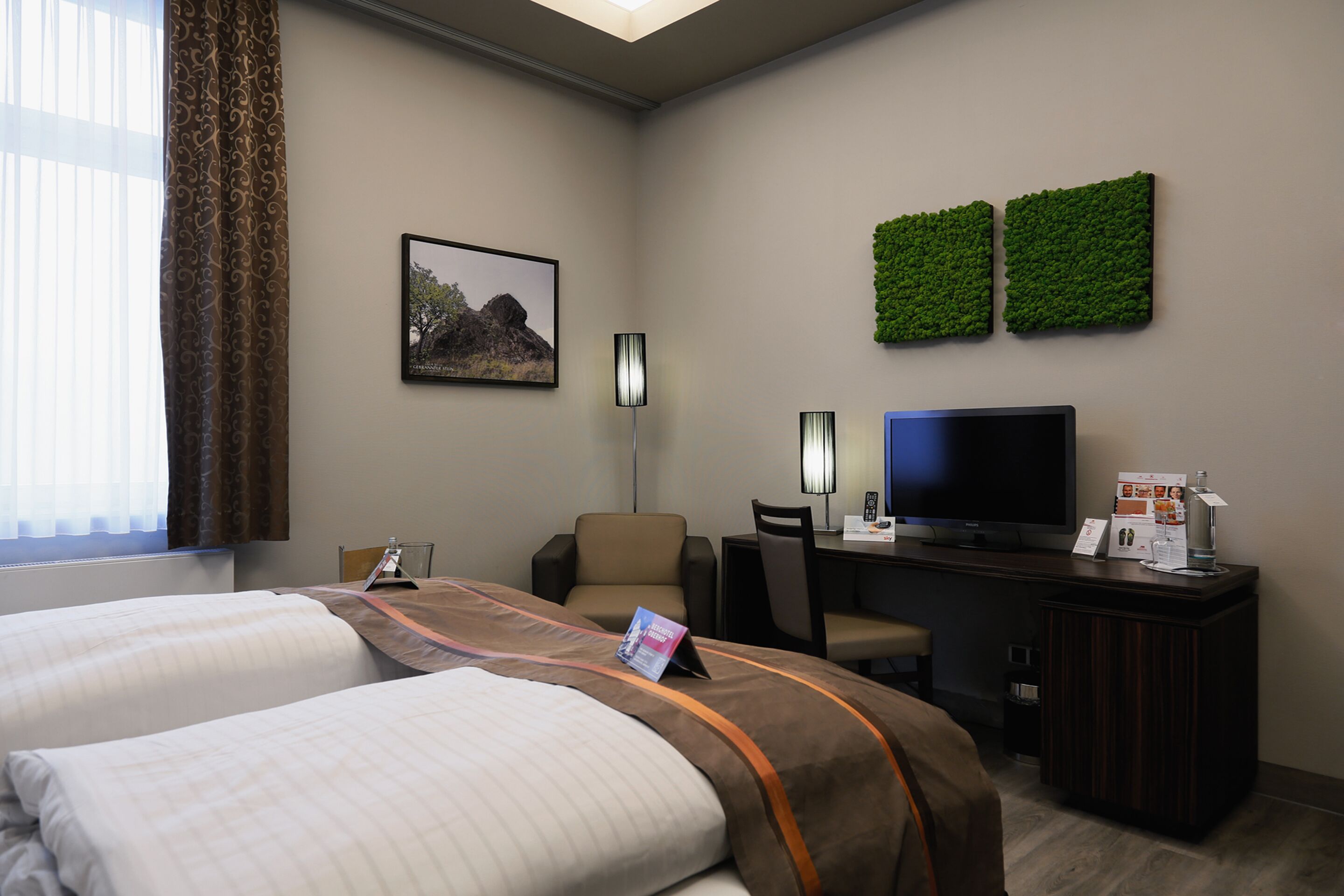 A hotel room with two beds, a desk with a computer, a TV, and a green wall decoration.