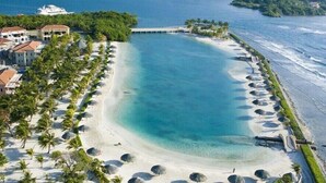 An aerial view of a beach resort with a large, clear blue pool surrounded by palm trees and a row of beach umbrellas. - Hotel Economico (Roatan)