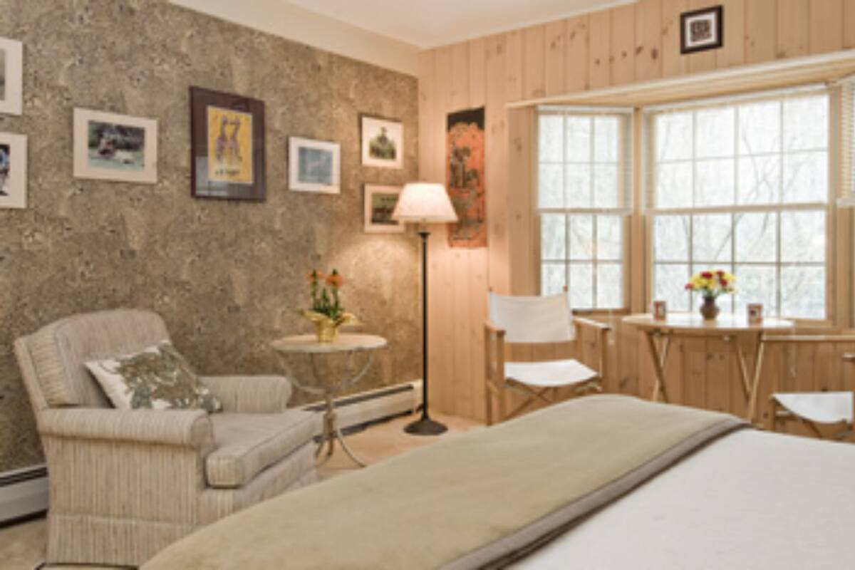 A room with a bed, a chair, a small table, and a lamp. There are framed pictures on the wall and a window with a view.