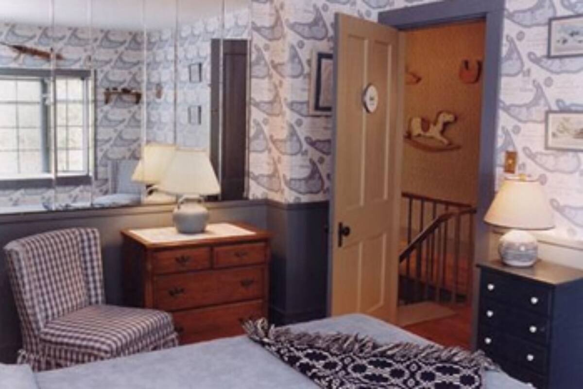 A bedroom with a checkered chair, a dresser, a bed with a patterned blanket, and a window with a view of the outside.