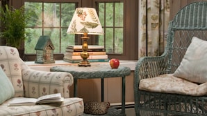 A cozy room with a floral-patterned sofa, a wicker chair, a small table with a lamp, and a bookshelf. - The Waterford Inne (Waterford)