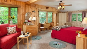 A cozy cabin-style room with a red sofa, a wooden desk, a bed with a red bedspread, and large windows offering a view of greenery. - The Waterford Inne (Waterford)