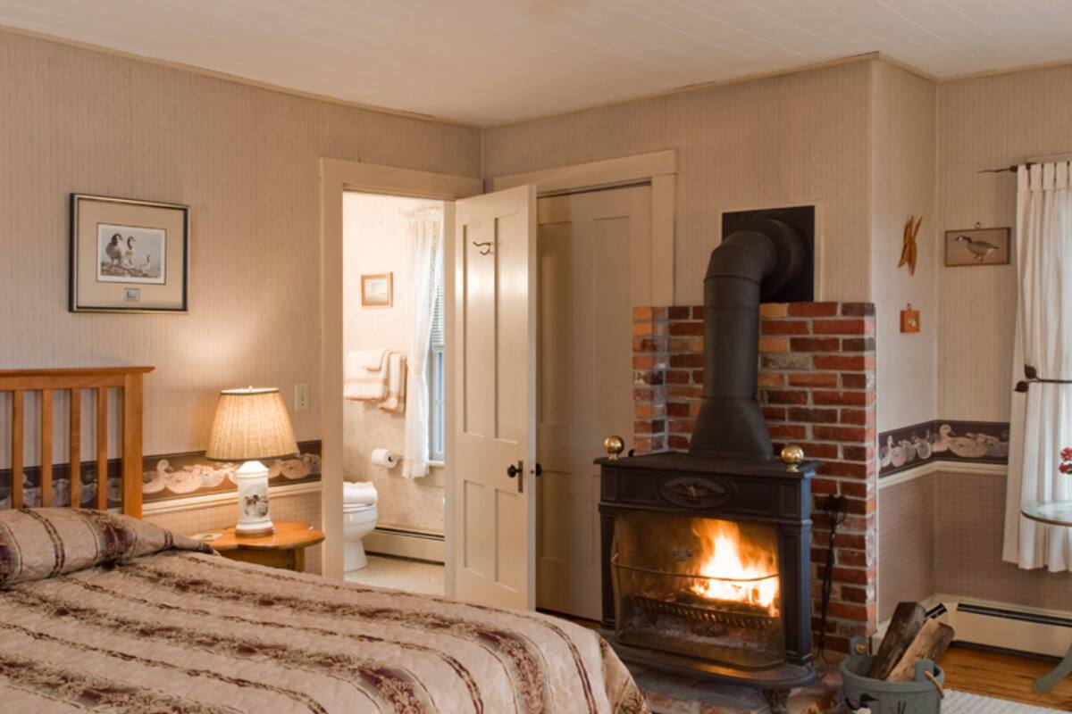 A cozy bedroom with a bed, a nightstand, a lamp, a fireplace, and a door leading to another room.