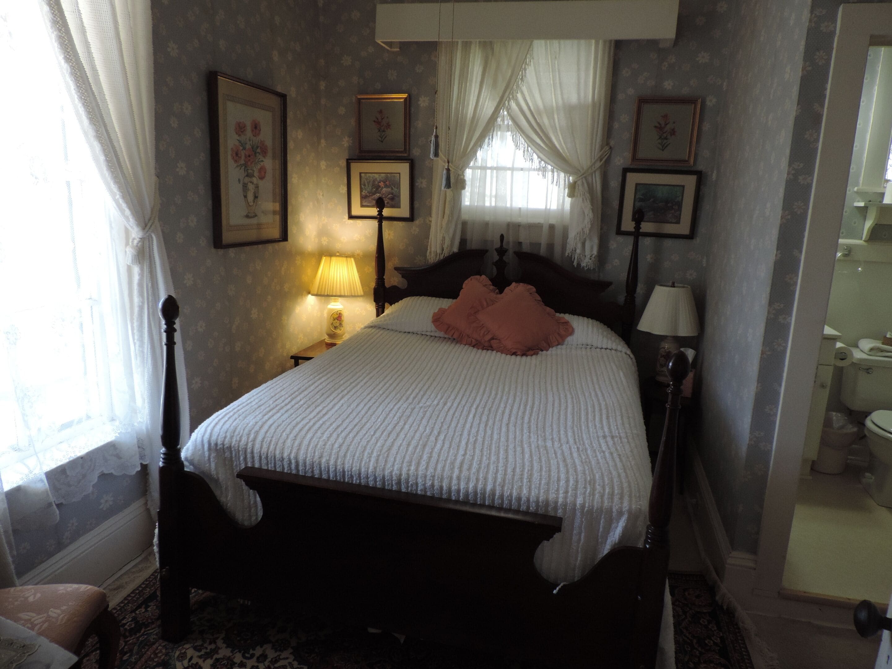 A bedroom with a bed, a nightstand with a lamp, a chair, and a bathroom visible through an open door.
