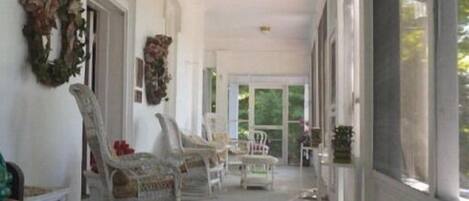 A covered porch with white wicker chairs, a dining table, and a large window providing a view of the outdoors.