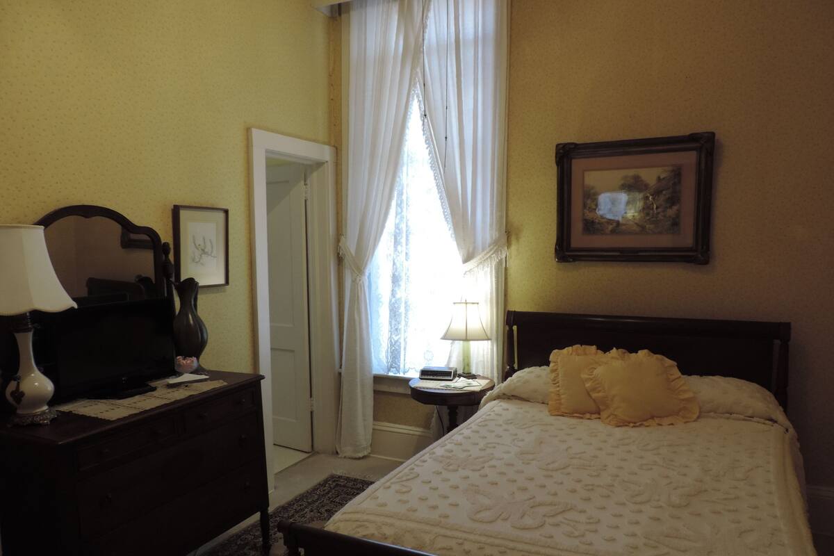 A bedroom with a bed, a bedside table, a lamp, a dresser, a mirror, a window with curtains, and a framed picture on the wall.