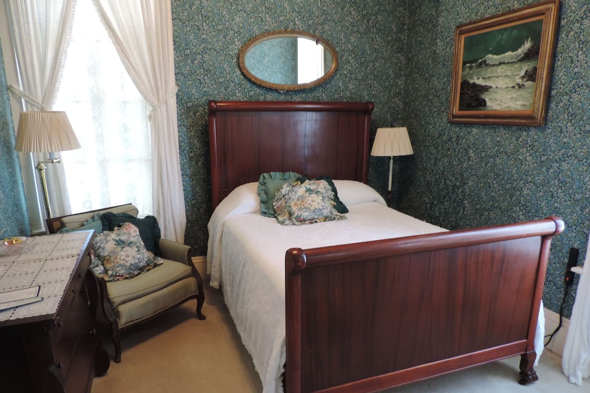 A bedroom with a wooden bed, a chair, a desk with papers, a lamp, a mirror, and a painting on the wall.