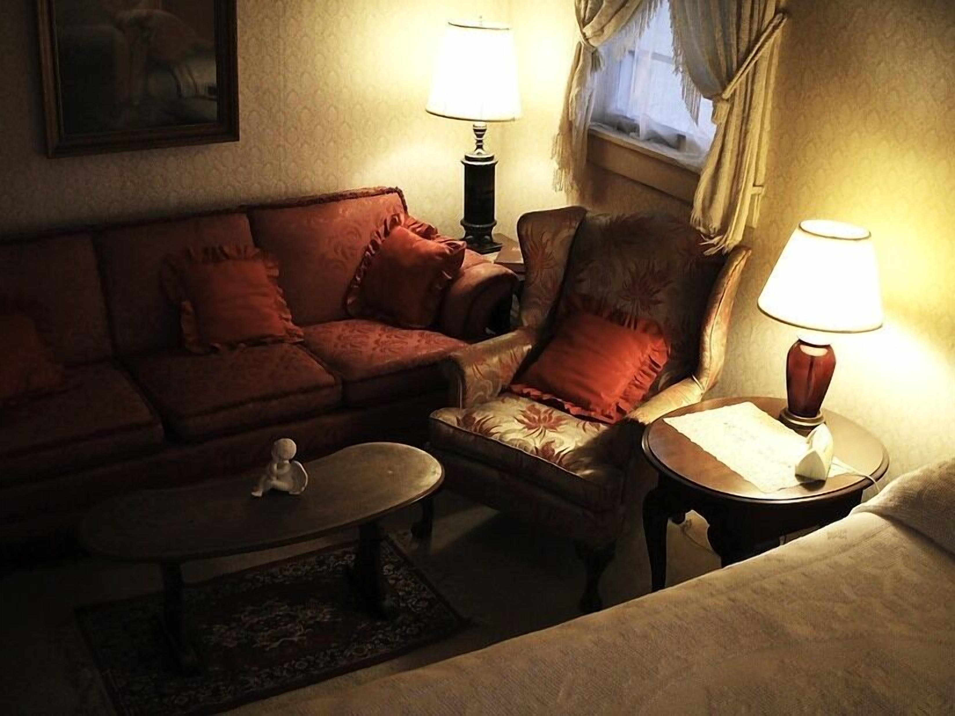 A cozy living room with a sofa, armchair, and a small table. There are two lamps on side tables and a window with curtains.