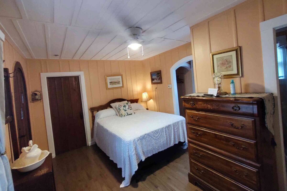 A bedroom with a wooden ceiling, a bed with a white bedspread, a wooden dresser, and a framed picture on the wall.