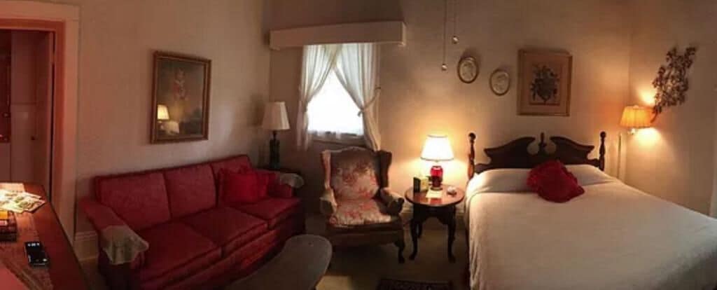 A hotel room with a bed, a red sofa, a chair, a bedside table with a lamp, and a window with curtains.