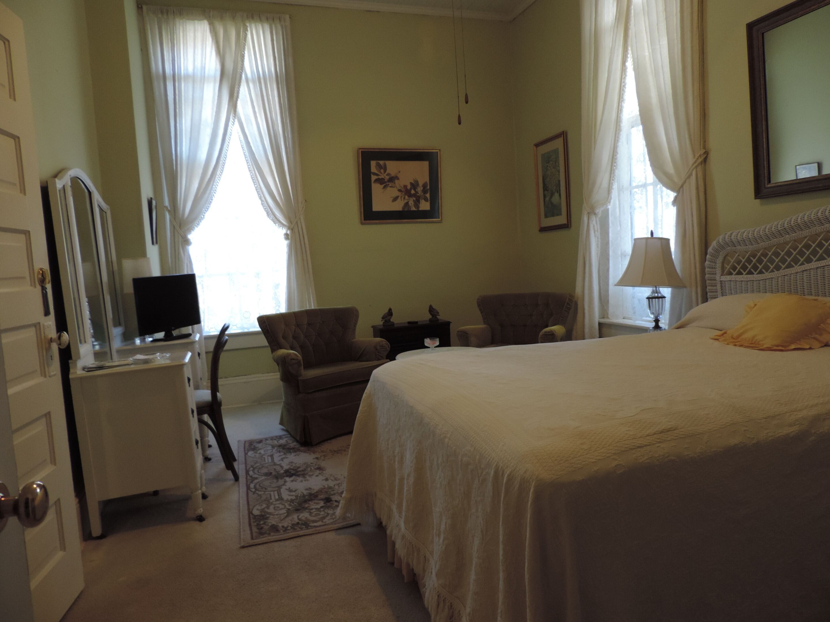 Une chambre à coucher comprenant un lit, deux fauteuils, un bureau avec un ordinateur, une lampe et une fenêtre avec des rideaux.