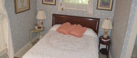 Un lit bien fait, recouvert d’une couvre-lit blanche et agrémenté d’oreillers roses, une table de chevet avec une lampe, et des décorations florales sur les murs.