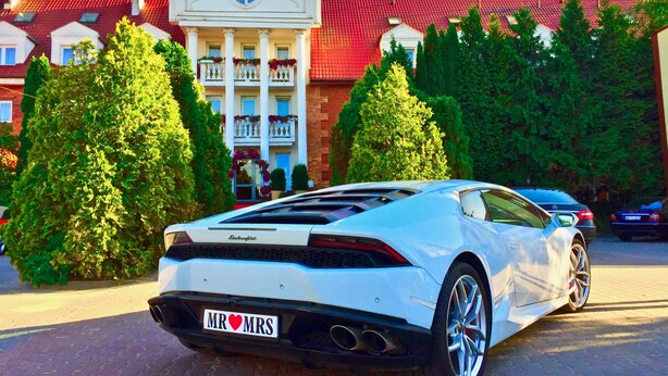 Un automóvil blanco de lujo estacionado frente a un hotel de estilo arquitectónico clásico, con un balcón central y techo rojo.