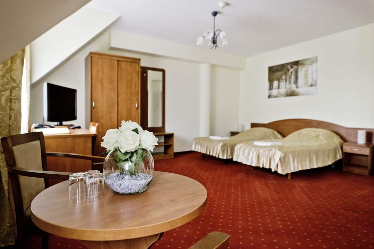 Una habitación de hotel con una mesa redonda de madera, dos sillas, una cama con edredón color beige, un armario de madera, un televisor y un candelabro.