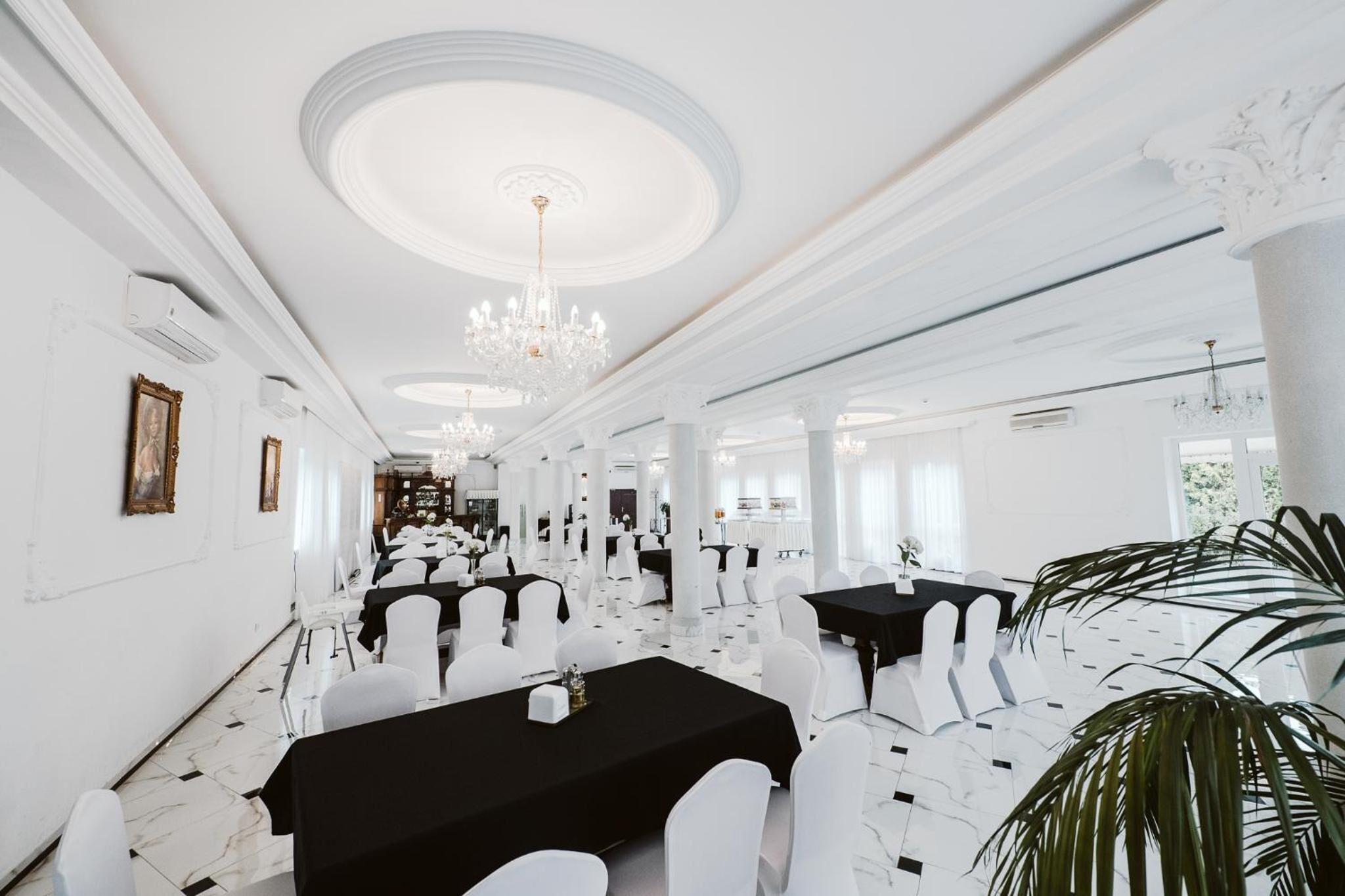 A spacious banquet hall with white walls, chandeliers, and a marble floor. Tables are set with white chairs and black tablecloths.